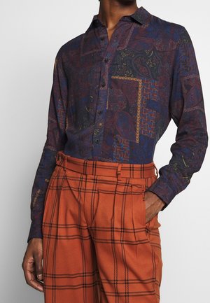 Homme portant une chemise boutonnée à motif sombre rentrée dans un pantalon orange taille haute avec des lignes à carreaux noires et une poche latérale.