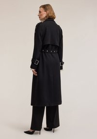 Czarny wełniany płaszcz z paskiem i srebrnymi elementami, z zapinanym na dwa rzędy guziki, naramiennikami i szerokimi mankietami. Elegancka, dopasowana sylwetka.