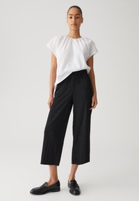 Witte korte mouwen top met geplooide halslijn, gecombineerd met zwarte wijde broek met een trekkoord. Zwarte instappers maken de look af.