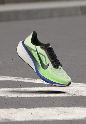 Zapatilla de correr Nike verde claro y negra con suela azul y blanca, suspendida en el aire sobre una superficie pavimentada con líneas blancas pintadas.