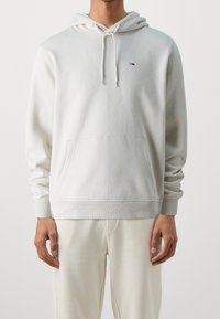 Vit huvtröja i mjukt tyg, med en framficka, ribbade ärmslut och en liten logotyp på bröstet. Stylad med ljusa byxor.