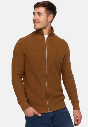 Suéter marrón de canalé con cremallera y cuello alto, que presenta mangas largas y un acabado texturizado. Llevado sobre una camisa blanca, combinado con vaqueros azules.