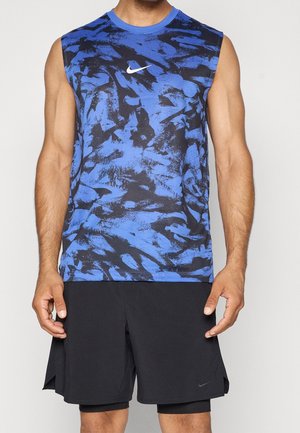 Mann trägt ärmelloses blau-schwarz abstrakt gemustertes Nike-Shirt und schwarze Nike-Shorts und steht vor einem einfarbigen Hintergrund.