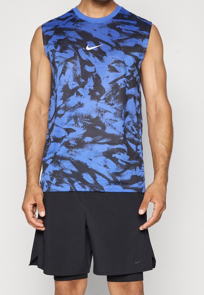 Homme portant un t-shirt Nike sans manches bleu et noir à motif abstrait et un short noir Nike, debout devant un fond uni.