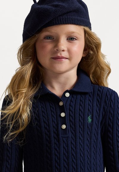 Mornarsko pulover s pletenim vzorcem in tremi belimi gumbi ter zelenim logotipom. Otrok nosi ujemajočo mornarsko pleteno beretko. Mehka tekstura in klasičen kroj.