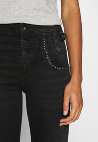 Jeans de mezclilla negra con cintura de tiro alto, cierre de botones y detalles decorativos de tachuelas en el bolsillo delantero. Textura suave y diseño ajustado.