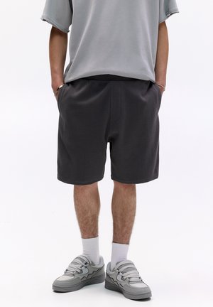 Dunkelgraue Shorts aus strukturiertem Stoff, mit einem elastischen Bund, seitlichen Taschen und lockerer Passform, kombiniert mit grauen Turnschuhen und weißen Socken.
