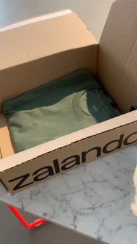 Pappkarp, millel on küljel trükitud "zalando", ja milles on volditud oliivroheline T-särk. Karp on lihtsa disainiga ja osaliselt avatud.