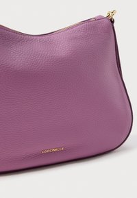 Lilla lærveske med struktur, gullglidelås og liten gull "Coccinelle"-logo foran, buet form.