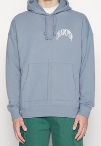 Světle modrá mikina s kapucí vyrobená z bavlny, s přední kapsou ve stylu kangaroo, žebrovanými manžetami a vytištěným logem "CHAMPION" v bílé barvě na hrudi.