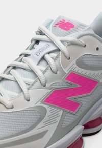 Chaussure de sport gris clair avec des accents roses, présentant une tige en mesh, des superpositions texturées et un design courbé, vue de côté et légèrement en hauteur.