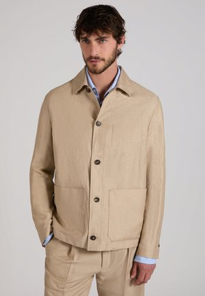 Mann trägt eine beigefarbene Jacke mit Knöpfen und passende Hose, hellblaues Hemd sichtbar am Kragen und an den Manschetten, steht vor schlichten Hintergrund.