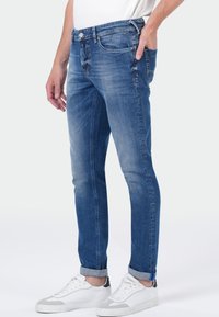 Homme portant un jean bleu slim avec des ourlets retroussés et des baskets blanches, se tenant avec une main dans la poche contre un fond uni.