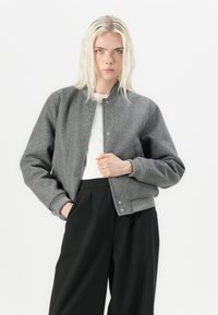 Giacca bomber grigia con una texture morbida, polsini a coste e chiusure a scatto. Abbinata a una maglietta bianca e pantaloni neri a gamba larga.