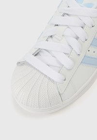 Ténis branco com detalhes azuis, biqueira texturada e atacadores planos. Parte superior sintética lisa e sola de borracha com padrão.