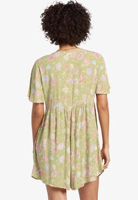 Kvinna med lockigt hår som bär en ljusgrön blommig klänning med korta ärmar, sedd bakifrån mot en vit bakgrund.