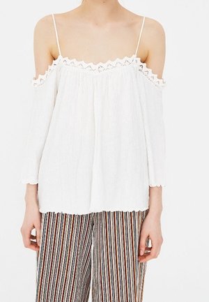 Blouse blanche à épaules dénudées avec finition en dentelle, tissu texturé et fines bretelles, assortie à un pantalon large rayé multicolore.