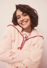 Roza jopa iz flisa z rdečimi izvezenimi srci in napisom ter z zadrgo. Ima visok ovratnik in mehak, teksturiran material. Nežen detajl z zadrgo.