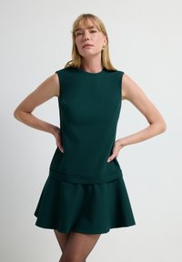 Ärmelloses Kleid aus tiefgrünem Stoff, tailliertes Oberteil mit ausgestellt Hem, glatte Textur und dezente Nahtdetails.