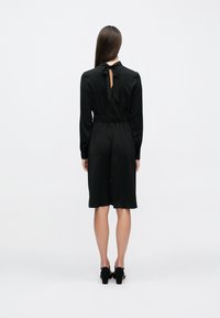 Rochie neagră din satin cu mâneci lungi, talie cambrată și fundă la gât. Partea din spate are o deschidere, iar tivul ajunge până la genunchi.