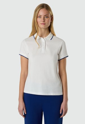 Donna con capelli biondi lisci che indossa una maglietta polo bianca con rifiniture blu e pantaloni blu, in piedi davanti a uno sfondo grigio chiaro uniforme.