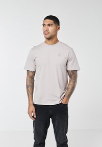 Bărbat cu păr negru scurt și brațe tatuate, purtând un tricou gri deschis și blugi negri, stând cu o mână în buzunar, pe un fundal alb.