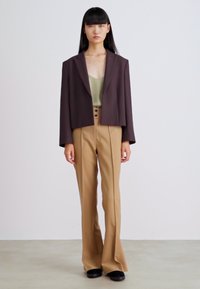 Mørkebrun cropped blazer over en lysegrønn topp, kombinert med beige utsvungne bukser og svarte sko. Jevne teksturer og tettsittende design.