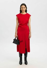 Eine Frau trägt ein ärmelloses rotes Midikleid mit Schlitz vorne, schwarze knielange Stiefel und hält eine kleine schwarze Handtasche vor einem schlichten Hintergrund.