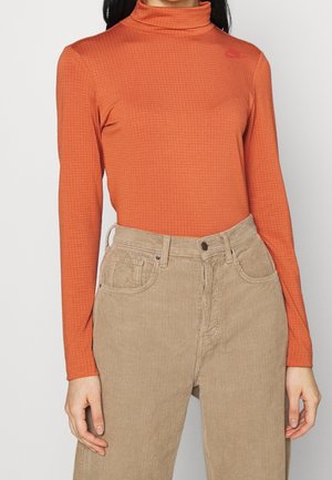 Frau trägt ein orangefarbenes, langärmliges Rollkragentop mit dezentem Karomuster und beige hoch taillierte Cordhosen.
