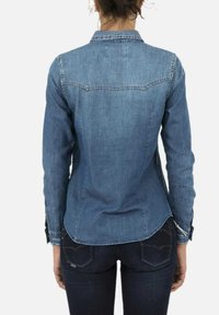 Camicia in denim azzurro chiaro a bottoni, con maniche lunghe e silhouette affusolata. Cuciture dettagliate lungo la spalla e i polsini.