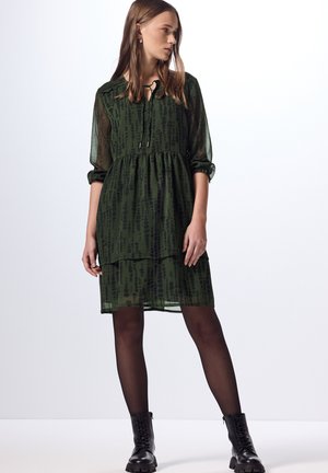 Robe verte à manches longues avec un design en couches, mettant en avant un col noué et un motif à pois noirs. Portée avec des bottes noires jusqu'aux genoux.