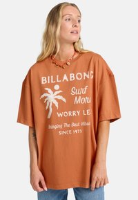 Camiseta de gran tamaño de color óxido, hecha de algodón, que presenta un gráfico de palmera blanca y un diseño de texto. Mangas cortas y corte relajado.