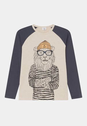 Beżowa i granatowa koszula z długim rękawem z ilustracją lwa w okularach, w paskowanej koszulce, pomarańczowej czapce typu beanie i z tatuażami na skrzyżowanych ramionach.