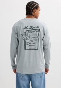 Camiseta de manga larga gris claro con un estampado gráfico negro en la parte posterior que incluye texto y una ilustración de un electricista. Corte casual y textura suave.