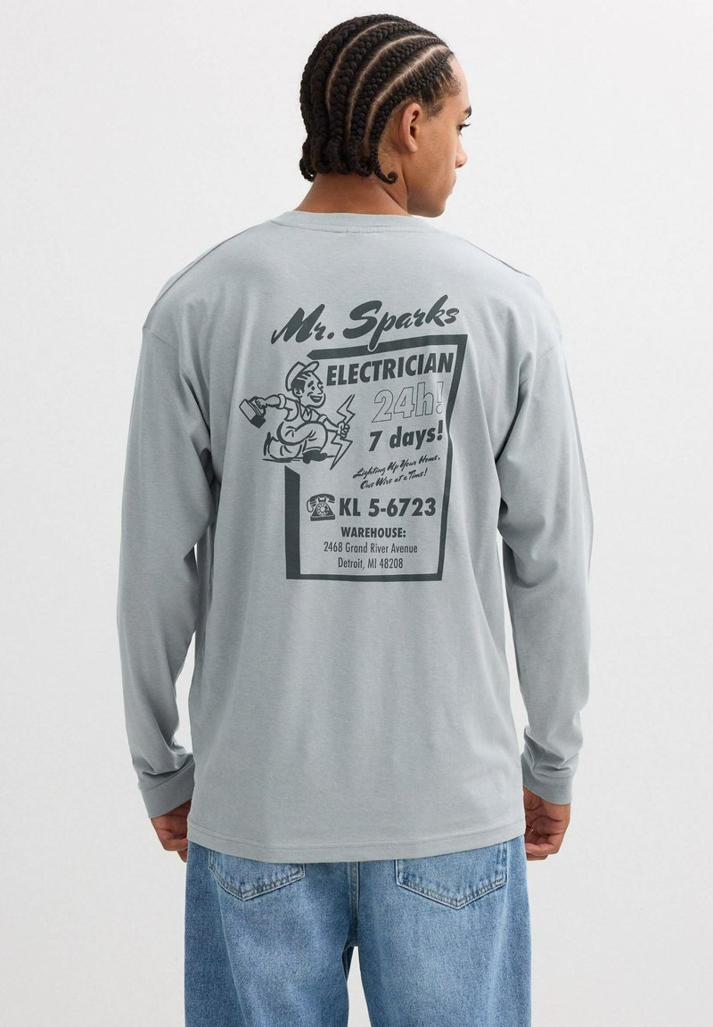 Camiseta de manga larga gris claro con un estampado gráfico negro en la parte posterior que incluye texto y una ilustración de un electricista. Corte casual y textura suave.
