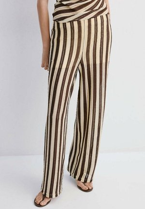 Osoba ubrana w brązowe i beżowe pionowo prążkowane dzianinowe spodnie z dopasowanym topem i czarne, odkryte sandały, stojąca na jasnej podłodze.