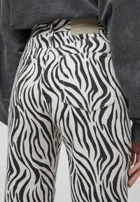 Jean en denim blanc avec un motif rayé zèbre noir, deux poches arrière et une étiquette sur la ceinture. La texture est lisse.