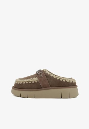 Chaussure slip-on en suède marron avec un accent tressé beige et un détail de boucle en argent, dotée d'une semelle beige épaisse et texturée.