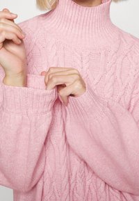 Pink knitted turtleneck sweater featuring cable knit pattern, ribbed neckline and cuffs, and slightly oversized sleeves for a relaxed fit.