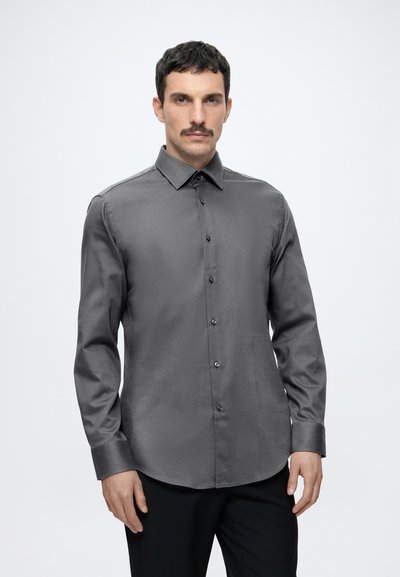 Homme aux cheveux foncés et moustache, portant une chemise grise à manches longues et un pantalon noir, debout sur un fond blanc uni.
