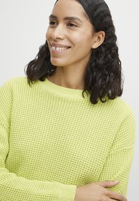 Limonengrüner Strickpullover mit Rundhalsausschnitt, strukturiertem Stoff, gerippten Bündchen und langen Ärmeln, getragen von einer lächelnden Frau mit lockigem Haar.