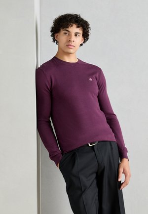 Langsleeve, ronde hals trui van bordeauxgebreid stof, met een klein logo op de borst. Gecombineerd met zwarte broek, model leunt tegen een muur.