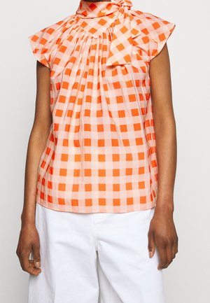 Mouwloze oranje-wit geruite blouse met een strik aan de hals, gedragen met witte broek met hoge taille.