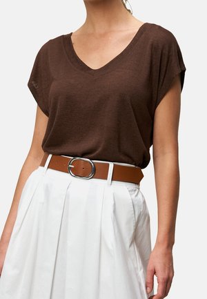Femme portant un haut marron à manches courtes et col en V rentré dans une jupe plissée blanche avec une ceinture en cuir marron et une boucle argentée.