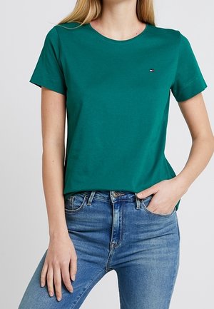 Kvinde iført teal kortærmet crew neck t-shirt med lille logo og blå denim jeans, hånd i lommen mod ensfarvet baggrund.