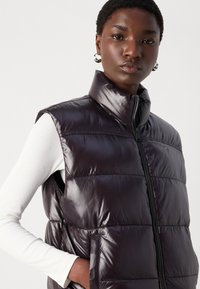 Sandro bez rukava, tamnoljubičasti prsluk od pufera s visokim ovratnikom i patentnim zatvaračem, s sjajnom završnom obradom i bočnim džepovima. Vidljiv je detalj teksturirane tkanine.