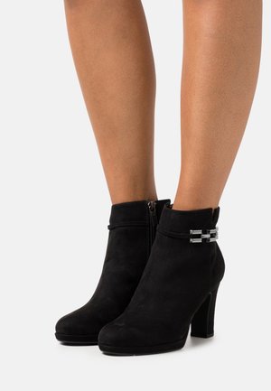 Bottines en suède noir avec une texture lisse, un haut talon bloc, une fermeture à zip et des accents métalliques décoratifs au niveau de la cheville.