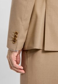 Beige jakkeslæb med fire brune knapper, twill tekstur og sidesplit. Nærbillede af en hånd, der hviler mod stoffet.