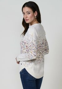 Pulover alb tricotat cu stropi multicolori, guler rotund și mâneci lungi. Prezintă un tiv asimetric și fante laterale.