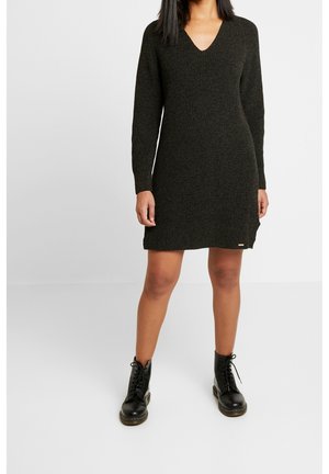 Kobieta w czarnej dzianinowej sukience na długi rękaw z dekoltem w kształcie litery V, w połączeniu z czarnymi sznurowanymi botkami do kostki, stojąca na jednolitym białym tle.